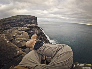 Tourist man sitting on a rock and looking at the endless sea. Wearing grey sport touristic pants, tan\beige army trekking boots.  Scenic waterfall in front of him, grey cliff and blue ocean on sides.