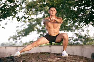 A muscular male athlete doing workout at the park. Gymnastics, training, fitness workout flexibility. Summer city in sunny day on background field. Active and healthy lifestyle, youth, bodybuilding.