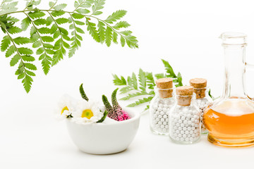 flowers in mortar near pestle and glass bottles with pills, jug with oil and green leaves on white