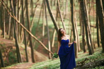 Beautiful girl in a blue dress walks in the woods with flowers