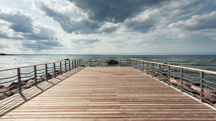 Fototapeta premium Perspective view at sea from center of wooden pier made of deck board with posts and ropes. Dramatic sky at daylight. Vintage toning