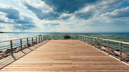 Fototapeta premium Perspective view at sea from center of wooden pier made of deck board with posts and ropes. Dramatic blue sky at beautiful sunny day