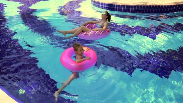 A happy young mother of European appearance, swims on an inflatable circle in the pool, watches her little daughter laughing and spinning merrily in the inflatable circle nearby.