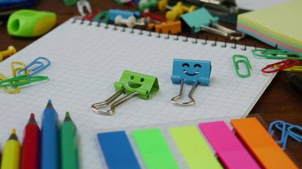 Smiles Blue and Green Binder Clips on Notebook with School Office Supplies: colored pencils and paper clips on brown wooden table. Concept of back to school and education - Powered by Adobe