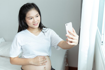 Cheerful pregnant young Asians woman sitting on bed and taking selfie with mobile phone at home.
