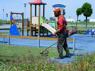Lawnmower Worker cuts grass with the trimmer near the playground