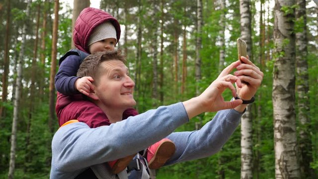 Man take selfie with baby girl sitting on his shoulders, happy two at walk in nature park. Father and little child looking at smartphone and smile