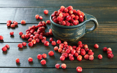 Wild strawberry in a clay cup on wooden dark brown background with forest berry scattering around, closeup, copy space, healthy food and vegetarian eating concept
