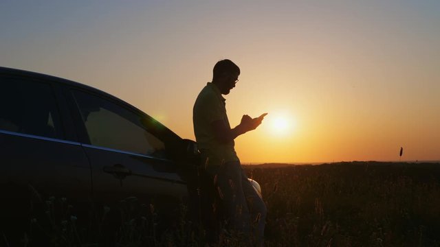 A man standing near a car uses a smartphone at sunset