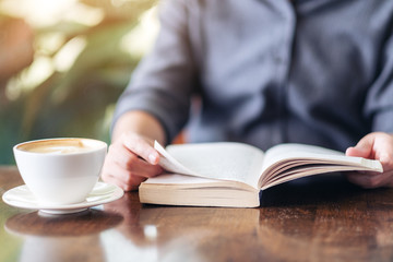 Closeup image of a woman holding and reading a book with coffee cup on wooden table