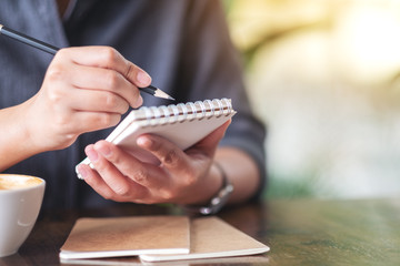 Closeup image of a woman holding and writing on blank notebook with coffee cup on table in cafe