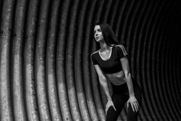 Black and white. Sports woman rest after run in fashion sportswear on tunnel urban background. Fitness model working out outdoor. Young beautiful slim brunette girl in trendy leggings and top.