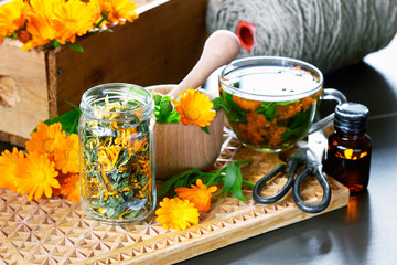 Calendula herb, a jar with dry marigold, a mortar with fresh bunch of flowers, a cup of herbal tea, a bottle of medicinal healing tincture and other stuff on wooden board,  closeup, copy space