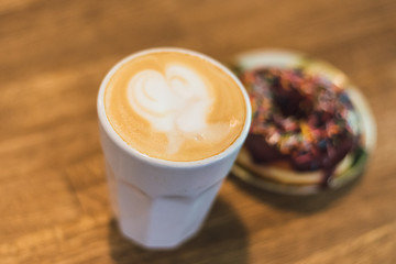 coffee with a heart made of milk and a donut on a wooden table. Coffee with a drawn heart and milk on a wooden table in a coffee shop. chocolate donut with scattering on the table next to the coffee