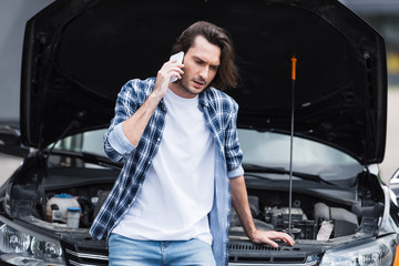 frustrated man talking on smartphone while standing near broken auto with open trunk, car insurance concept