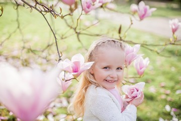 Fototapeta premium Little cute blonde girl 3 years old plays in a park near a blossoming magnolia.