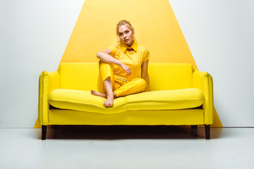 Fototapeta na wymiar blonde girl with barefoot sitting on sofa and looking at camera on white and yellow