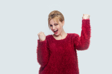 Portrait of happy successful rejoicing beautiful young blond woman in red blouse standing, and celebrating her victory with surprised amazed face. indoor studio shot isolated on light gray background.