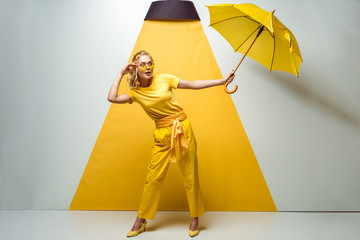 attractive blonde girl touching sunglasses and holding umbrella on white and yellow