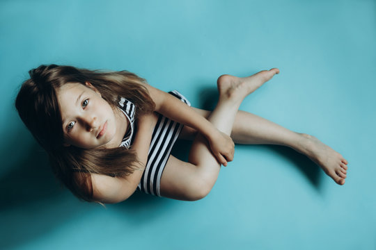 Girl sitting on plain blue background looking at camera. Child glancing up top view. Simple wallpaper, background. Serious and sad facial expression. Kid in striped dress with legs crossed