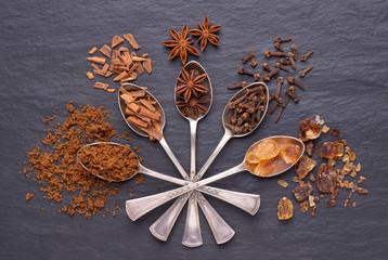 Aromatic spices and brown sugar in silver spoons on black stone background