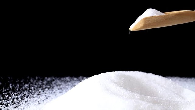 White granulated sugar on wooden spoon pouring growing into pile against on black background.