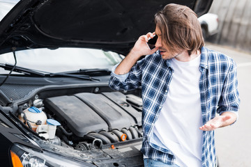 upset man in casual wear talking on smartphone near broken auto with open trunk, car insurance...