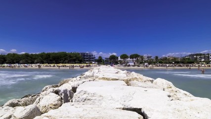 Timelapse of a pier and the beach from which it is born, Alba Adriatica, Teramo, Abruzzo. The pier is made of large white stones and beyond the beach you can see buildings and trees.