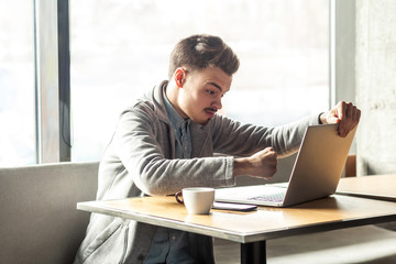 Side view portrait of aggressive unhappy young businessman in grey blazer are sitting in cafe and having bad mood are ready to punch a worker through a webcam with raised fist with angry face. indoor