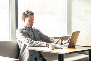 Stop! Side view portrait of emotional scared young businessman in grey blazer are sitting in cafe and screaming to the laptop cause made big mistake with raised arms and stressed face. Indoor