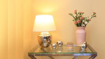 Lampka nocna i kwiaty w wazonie na szklanym stoliku i złotych barwach.	