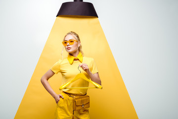 attractive woman in sunglasses holding hanger and standing with hand in pocket on white and yellow - obrazy, fototapety, plakaty