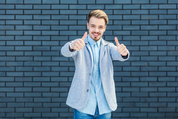 Portrait of happy handsome young blonde man in casual style standing with thumbs up, like gesture, and looking at camera with toothy smile. indoor studio shot on brick wall background.