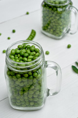 Glass jars with fresh green peas on white wooden background, copy space. Bio healthy food. Pods and pea leaves on wood table