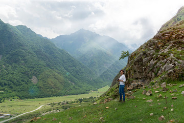 Fototapeta na wymiar young woman in a white shirt and jeans stands against the mountain and looks at the valley. Photographer examines the landscape. Toned