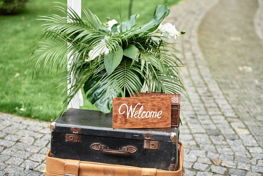 Wedding decorations with welcome sign board, flowers, pulm leaves and vintage suitcases in garden, copy space