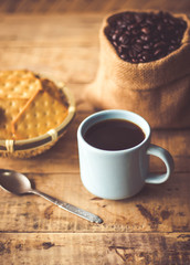 Black coffee In the cup sky blue color have roasted coffee beans are In a cloth bag sack butter cracker In the basket weave bamboo, and still have stainless steel spoon all placed on a wooden table.