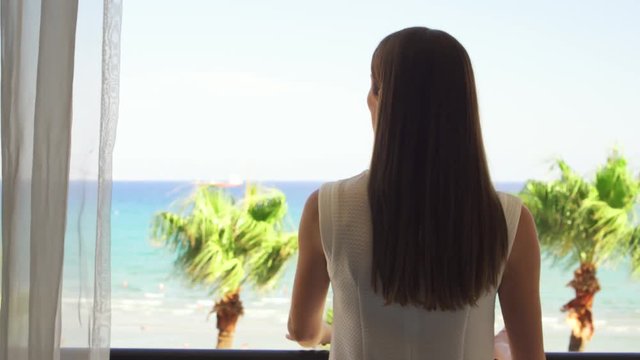 Young woman going out onto balcony and enjoying view from terrace. Female on vacation opening curtains and looking at Mediterranean sea. Wind swaying leaves on palm trees. Camera follows.