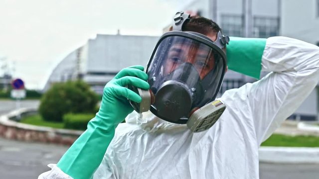 Portrait of hurrying young man in protective costume wearing gas mask against a power station at daytime. Fearful man suffocating by poisonous air in exclusion radiation zone in Chernobyl. Apocalypse.