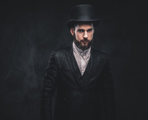 Portrait of a stylish bearded male in an elegant suit and cylinder hat, looking at a camera over dark background.