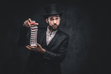 A Bearded magician in a black suit and top hat, showing trick with playing cards on a dark...