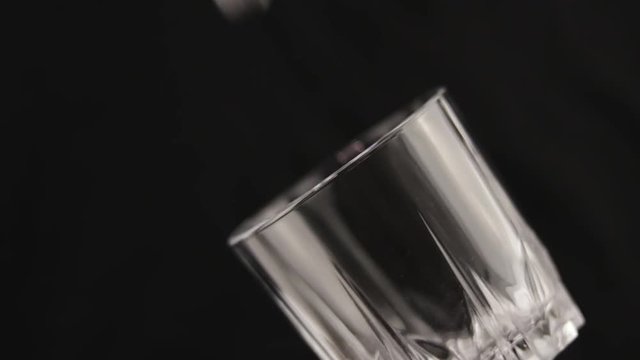 Ice cubes slowly fall into a glass cup for whiskey which is angled on a black background