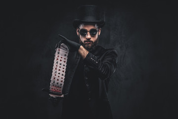 Magician in a black suit, sunglasses and top hat, showing trick with playing cards on a dark...