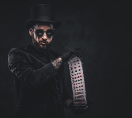 Magician in a black suit, sunglasses and top hat, showing trick with playing cards on a dark...