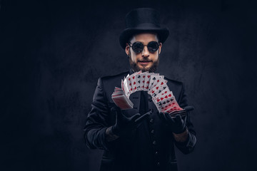 Magician in a black suit, sunglasses and top hat, showing trick with playing cards on a dark...