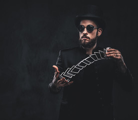 Magician in a black suit, sunglasses and top hat, showing trick with playing cards on a dark...