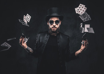 Magician in a black suit, sunglasses and top hat, showing trick with playing cards on a dark...