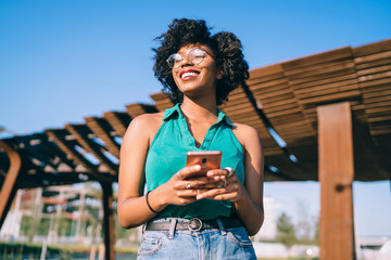 Below view of excited female blogger in stylish eyewear using modern technology and enjoying weather on sunny summer day, happy dark skinned hipster girl holding mobile phone, millennial generation