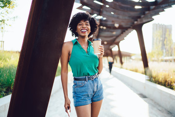 Half length portrait of cheerful dark skinned female with takeaway coffee cup posing for camera in city at summer, carefree andhappy African American hipster girl in casual wear enjoying sunny weather