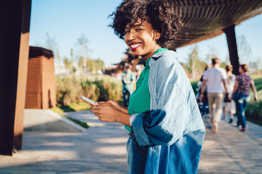 Half length portrait of cheerful dark skinned female feeling excited using 4G LTE internet connection on cellphone,positive smiling hipster girl dressed in stylish causal apparel ckeck online banking